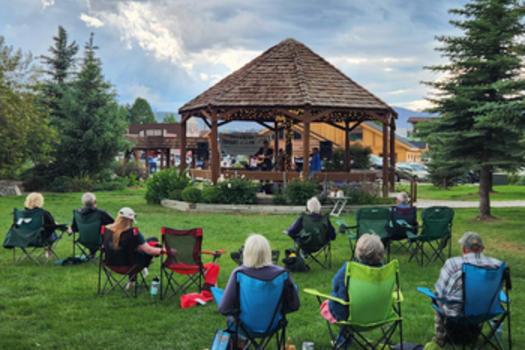 People sitting in camp chairs on a lush green lawn watching a concert in the Town Park gazebo